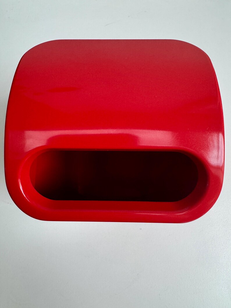 Puede incluir: Un contenedor rectangular rojo brillante con una parte superior redondeada y una gran abertura ovalada. El contenedor tiene un acabado liso y brillante y parece estar hecho de pl&aacute;stico. La abertura revela un interior oscuro.
