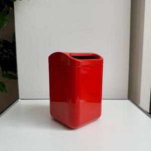Puede incluir: Un cubo de basura rojo brillante, rectangular, con la parte superior curvada y el interior negro. El cubo de basura est&aacute; hecho de un material brillante y se encuentra sobre una superficie blanca. El dise&ntilde;o es moderno y minimalista.