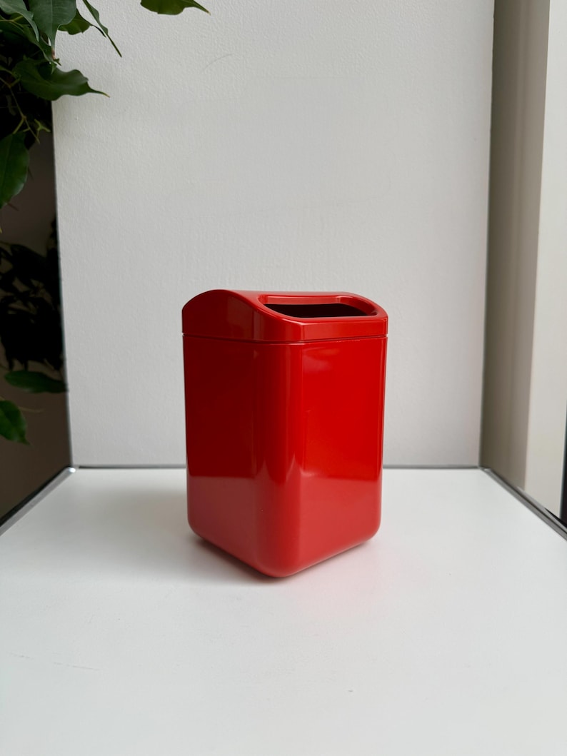 Puede incluir: Un cubo de basura rojo brillante, rectangular, con la parte superior curvada y el interior negro. El cubo de basura est&aacute; hecho de un material brillante y se encuentra sobre una superficie blanca. El dise&ntilde;o es moderno y minimalista.