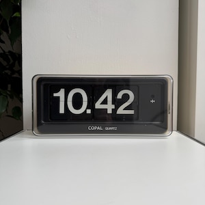 May include: A black and silver Copal Quartz flip clock displaying the time 10:42. The clock has a rectangular shape with a clear plastic cover. The clock is on a white surface.