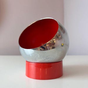 May include: A chrome-finished, spherical object with a red interior, resting on a cylindrical red base. The top of the sphere is cut away, revealing the red interior. The design is modern and minimalist.