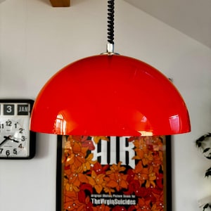 May include: A bright red, domed pendant light. The light is suspended by a black, coiled cable. A framed poster with floral artwork and the text "AIR" and "The Virgin Suicides" is visible in the background.