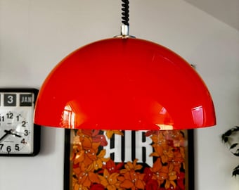 May include: A vibrant red, dome-shaped pendant light fixture. The light hangs from a black, coiled cord. A framed poster with floral imagery and the text "AIR" and "The Virgin Suicides" is visible in the background.