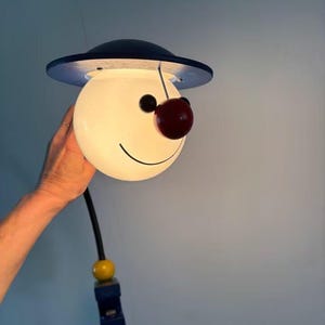 May include: A whimsical, smiling lamp with a white globe-shaped head, a black hat, and a cherry-red nose. The lamp has a black cord and a yellow and blue base. The lamp is held by a hand against a light blue background.