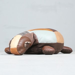 Op de afbeelding: Een patchwork leren ottomane in tinten bruin, beige en blauw. De ottomane is gevormd als een schildpad en heeft een zachte, pluche textuur.