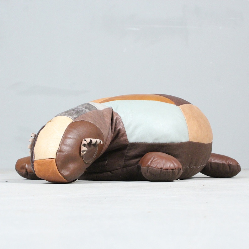 May include: A patchwork leather ottoman in the shape of a badger. The ottoman features a variety of brown, tan, and cream-colored leather patches. The badger has small, rounded ears and four stubby legs. The ottoman is designed to be a decorative and functional piece of furniture.