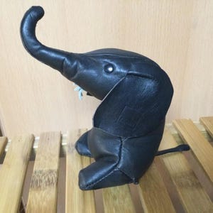May include: A black leather elephant figurine with a long trunk and a textured surface. The elephant is sitting on a wooden surface.