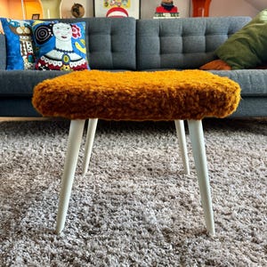 May include: A small, mid-century style stool with a rectangular, mustard-yellow, textured seat. The stool has four white, tapered legs. The stool is placed on a grey shag rug in front of a blue sofa.