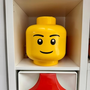 May include: A large, yellow storage container shaped like a Lego figure's head. The container has black eyes and a curved black smile. It is sitting on a white shelf inside a white cabinet.
