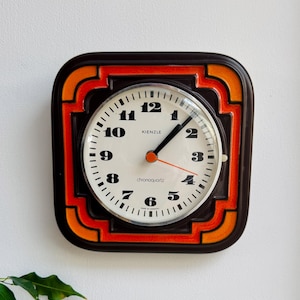May include: A square Kienzle wall clock with a dark brown and orange ceramic frame. The circular face is off-white with black numerals from 1 to 12, black hour and minute hands, and a thin orange second hand. The text Kienzle and chronoquartz are visible.