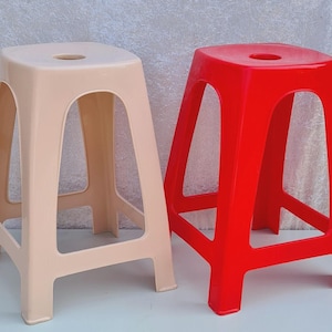 May include: Two plastic stools, one beige and one red, are displayed side-by-side. Each stool has a square seat with a circular hole in the center and four angled legs. The stools are made of a smooth, solid plastic material.