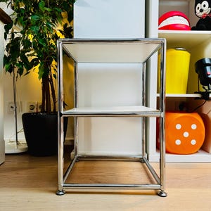 May include: A chrome metal and white shelf unit with two levels. The unit has a rectangular frame and sits on four small, round feet. The shelves are white. A potted plant is visible in the background.