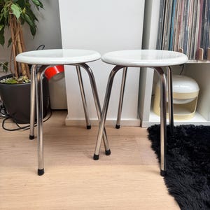 May include: Two white metal stools with chrome legs and black rubber feet. The stools are on a light wood floor.