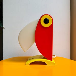 Könnte beinhalten: Eine toucanförmige Lampe mit rotem Kopf, gelbem Auge und weißem Schnabel. Die Lampe steht auf einem gelben Sockel und hat ein weißes Kabel. Die Lampe ist ein dekorativer Artikel.