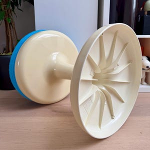 May include: A vintage, off-white and blue cleaning tool with a round, sponge-like pad. The tool has a stem connecting the pad to a circular base. The base is off-white with a fan-like design on the underside. The tool is resting on a wooden surface.