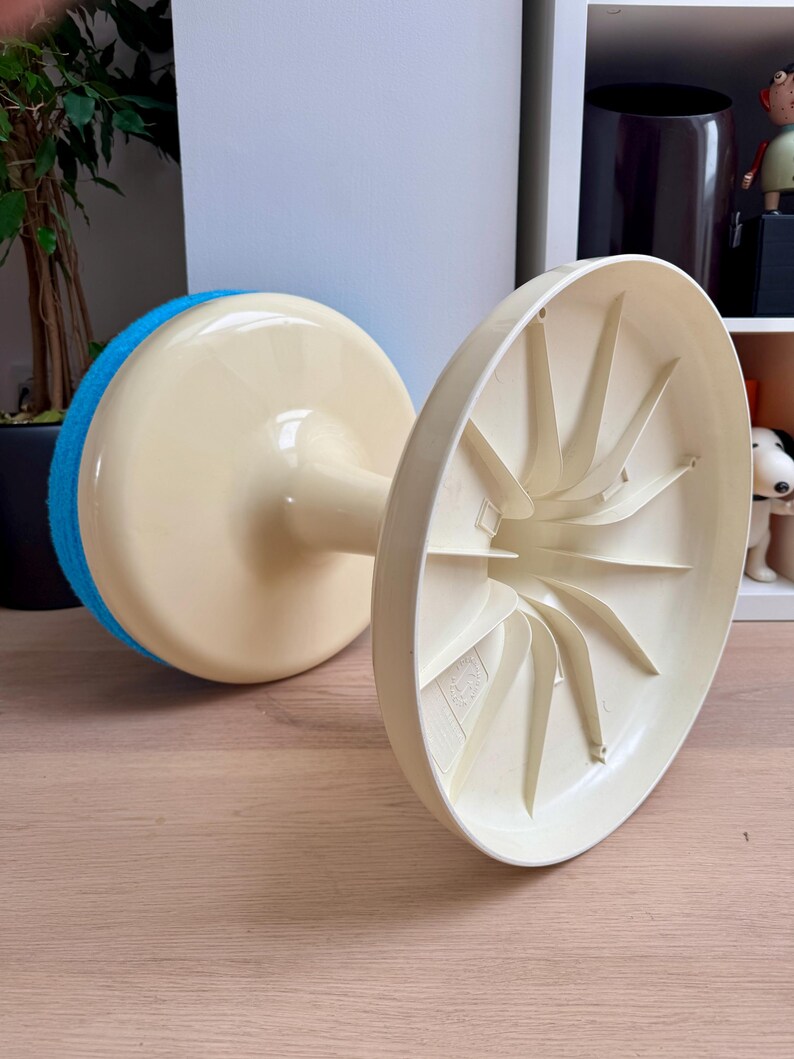 May include: A vintage, off-white and blue cleaning tool with a round, sponge-like pad. The tool has a stem connecting the pad to a circular base. The base is off-white with a fan-like design on the underside. The tool is resting on a wooden surface.