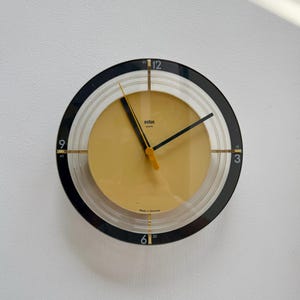 Puede incluir: Un reloj de pared redondo de cuarzo Braun con esfera central amarilla, manecillas negras y segundero amarillo. El reloj presenta un anillo exterior negro con los números blancos 12, 3, 6 y 9, y un marco transparente. El texto "Braun" y "Made in Germany" está impreso en la esfera.
