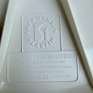 May include: Close-up of a white plastic object with embossed text. The text includes the name "LOCKHAUSEN PLASTIK" in a circular design and "FRIEDRICH STUCKENBR&Ouml;KER" with additional details about the manufacturer.