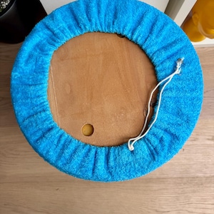 May include: A round, wooden stool with a bright blue terry cloth cover. The cover is gathered around the edge and has a white drawstring. The stool is viewed from above, showing the wooden top and the blue cover.