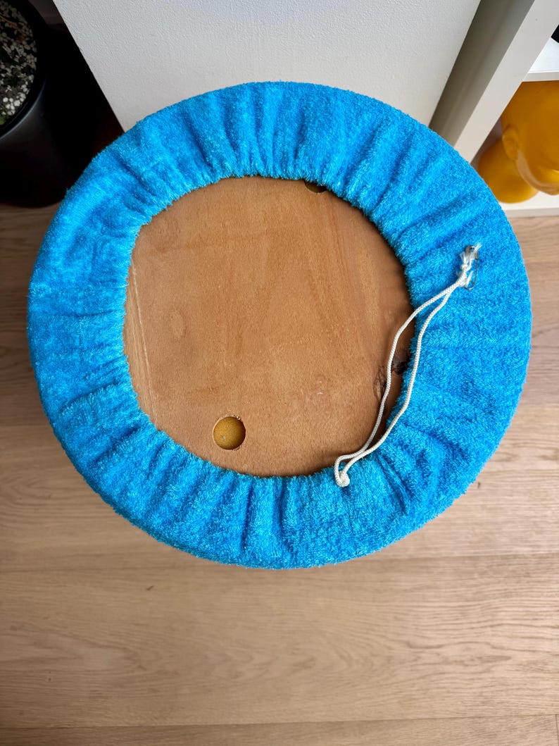 May include: A round, wooden stool with a bright blue terry cloth cover. The cover is gathered around the edge and has a white drawstring. The stool is viewed from above, showing the wooden top and the blue cover.