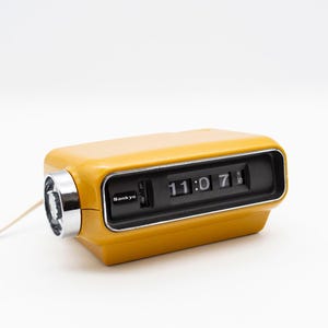 Peut inclure: Une horloge à bascule Sankyo jaune vintage affichant 11h07. L'horloge est de forme rectangulaire avec un cadran noir et des détails chromés. La marque "Sankyo" est visible. Un cordon blanc sort du côté.