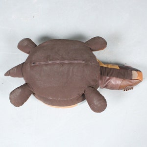 May include: A brown turtle-shaped pillow with a zipper closure on its back. The pillow features a mix of brown fabrics and leather, with a lighter brown underside and a tan and orange head.
