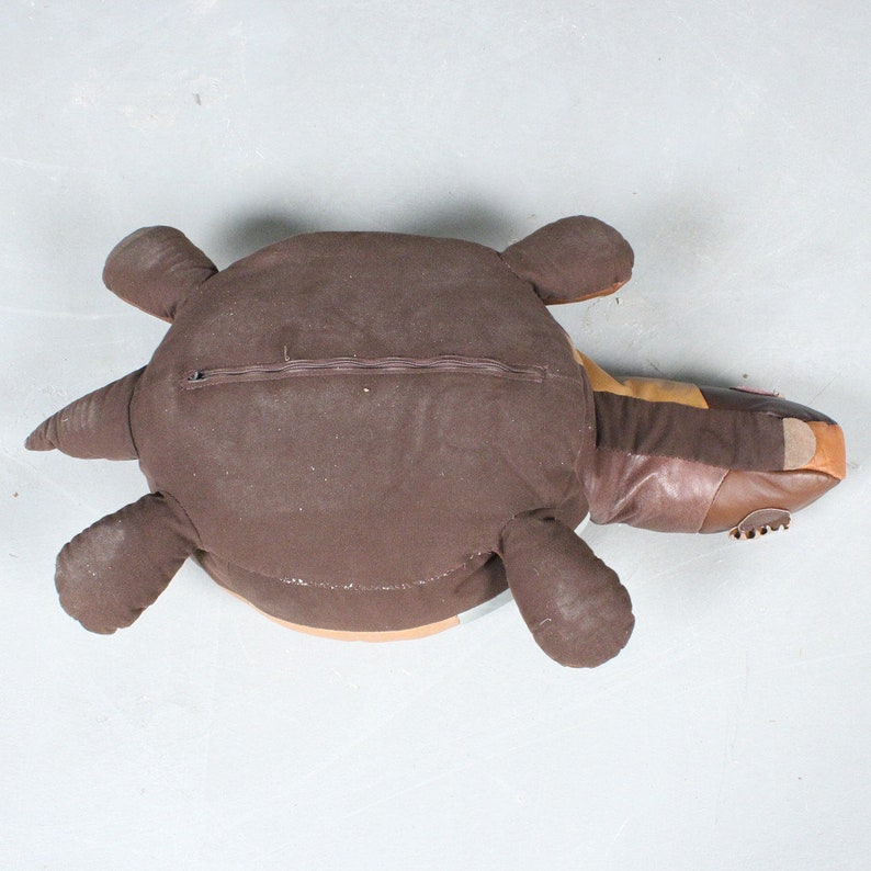 May include: A brown turtle-shaped pillow with a zipper closure on its back. The pillow features a mix of brown fabrics and leather, with a lighter brown underside and a tan and orange head.