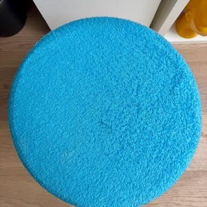 May include: A round, bright turquoise rug with a soft, textured surface. The rug is circular and appears to be made of a plush material. The rug is placed on a light-colored wooden floor.