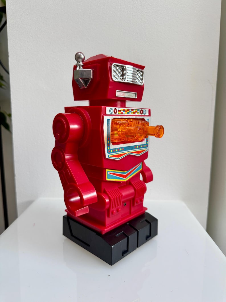 May include: A red toy robot with a silver antenna, rectangular eyes, and a clear orange chest piece. The robot has a black base and articulated arms. A retro-style toy.