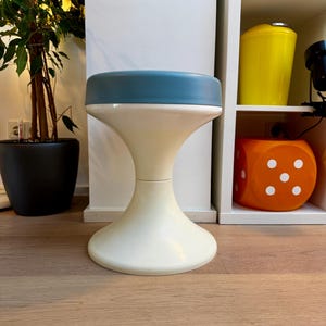 Pode incluir: Um banco retro com base branca e assento azul-petróleo. O banco tem formato de ampulheta, com uma base larga que se estreita antes de se alargar novamente para suportar o assento. O design lembra os móveis modernos de meados do século. Altura: aprox. 45 cm.