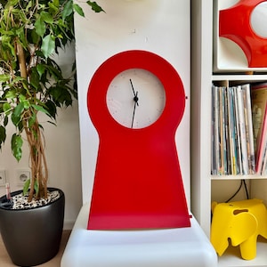 Pode incluir: Um relógio vermelho brilhante em forma de buraco de fechadura, com um mostrador branco e ponteiros pretos. O relógio está sobre uma superfície branca. O fundo inclui uma planta em vaso e uma prateleira com objetos decorativos. O design do relógio é moderno e minimalista.