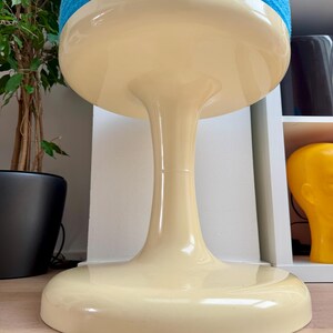 May include: A retro-style stool with a cream-colored base and a round, blue, textured seat. The stool has a unique, hourglass-shaped stem. The base is wide and flat, providing stability. The overall design is reminiscent of mid-century modern furniture.