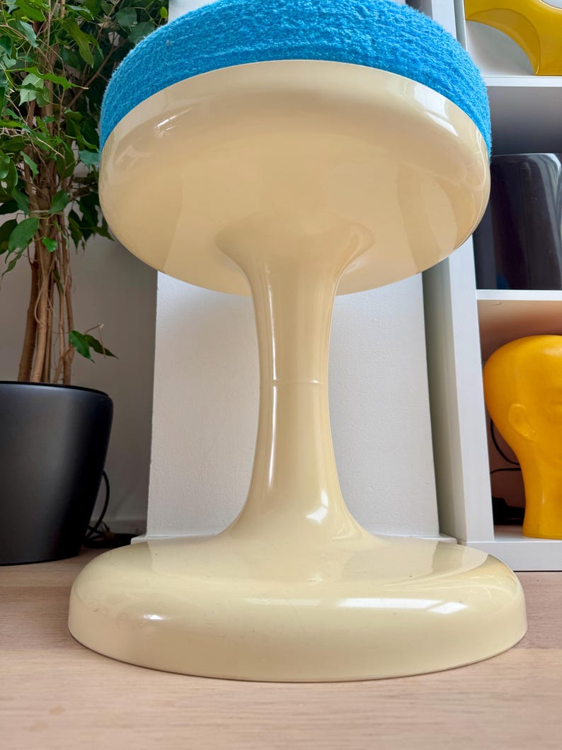 May include: A retro-style stool with a cream-colored base and a round, blue, textured seat. The stool has a unique, hourglass-shaped stem. The base is wide and flat, providing stability. The overall design is reminiscent of mid-century modern furniture.