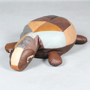 May include: A patchwork leather turtle-shaped ottoman in shades of brown, tan, and gray. The turtle has a rounded shell, a long neck, and four stubby legs. The ottoman is designed for use as a footrest or decorative piece.