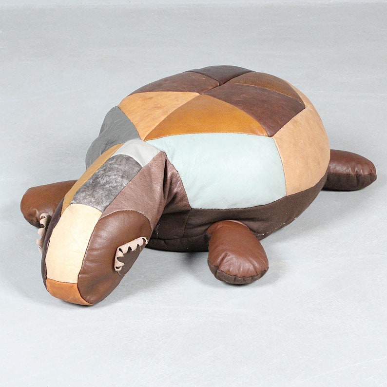 May include: A patchwork leather turtle-shaped ottoman in shades of brown, tan, and gray. The turtle has a rounded shell, a long neck, and four stubby legs. The ottoman is designed for use as a footrest or decorative piece.