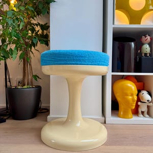 May include: A retro-style stool with a cream-colored base and stem, topped with a round, bright blue, textured seat. The stool has a wide, circular base and a flared stem, creating a unique, eye-catching design. The stool is a decorative furniture piece.