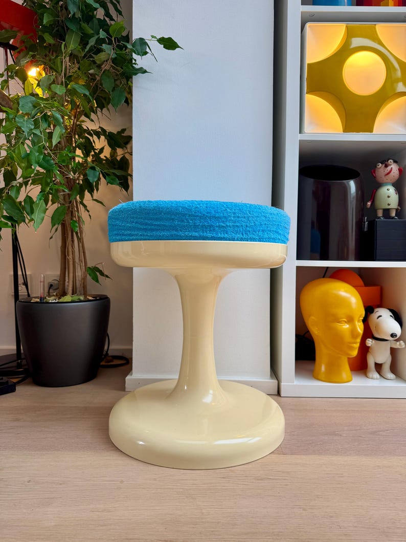 May include: A retro-style stool with a cream-colored base and stem, topped with a round, bright blue, textured seat. The stool has a wide, circular base and a flared stem, creating a unique, eye-catching design. The stool is a decorative furniture piece.