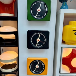 Op de afbeelding: Drie vierkante klokken in groen, zwart en geel. Elke klok heeft een zwarte wijzerplaat met witte markeringen en wijzers, en een rode secondewijzer. De klokken zijn op een witte muur gemonteerd.