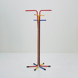 May include: A colorful coat rack with a minimalist design. The rack features a tall, vertical pole in yellow, blue, and red, with horizontal arms at the top and base. The arms are red, yellow, and blue. The rack is made of metal.