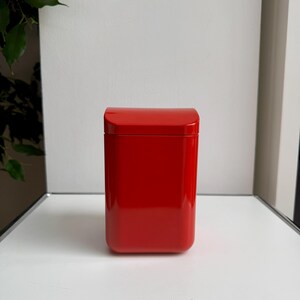 Puede incluir: Una caja roja rectangular y brillante con una parte superior curvada. La caja es de un color rojo s&oacute;lido y vibrante y se encuentra sobre una superficie blanca. El fondo es blanco neutro, con un toque de follaje verde visible a la izquierda.