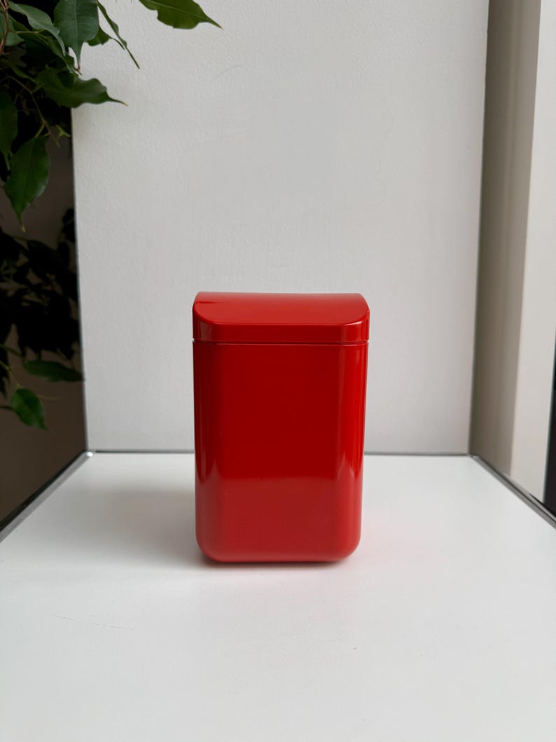 Puede incluir: Una caja roja rectangular y brillante con una parte superior curvada. La caja es de un color rojo s&oacute;lido y vibrante y se encuentra sobre una superficie blanca. El fondo es blanco neutro, con un toque de follaje verde visible a la izquierda.