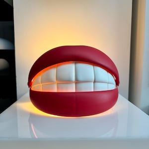 May include: A novelty lamp shaped like a smiling mouth. The lamp features bright white teeth, red lips, and an internal light source that illuminates the teeth. The lamp is sitting on a white surface.