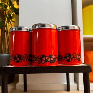 Puede incluir: Tres botes rojos con tapas plateadas. Cada bote tiene un diseño floral en negro, rosa y azul cerca de la base. Los botes cilíndricos están sobre una pequeña mesa negra.