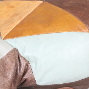 May include: Close-up of a patchwork leather pouf. The pouf features a geometric design with various leather pieces in shades of brown, tan, and off-white. The leather has a smooth texture and is stitched together to form a rounded shape.