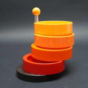 Peut inclure: Un organisateur de bureau en plastique empilable multicolore avec un bouton rond orange sur le dessus. L'organisateur est composé de quatre anneaux empilés dans des tons d'orange et de rouge.