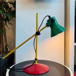 May include: A retro-style desk lamp with a green conical shade, yellow arm, and red base. The lamp has a black cord and a black adjustable joint. The lamp is on a black and white table.