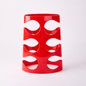 May include: Red plastic wine bottle holder with a modern design. The holder has six slots for bottles and is shaped like a cylinder.