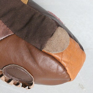 May include: Close-up of a handcrafted leather shoe with a patchwork design. The shoe features various shades of brown leather, including dark brown, tan, and a lighter brown suede accent. The design includes a stitched edge and a decorative leather detail.