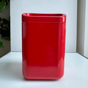 Puede incluir: Un recipiente rojo brillante de forma rectangular con una parte superior ligeramente empotrada. El recipiente tiene esquinas redondeadas y una superficie lisa y reflectante. El fondo es una superficie blanca lisa.