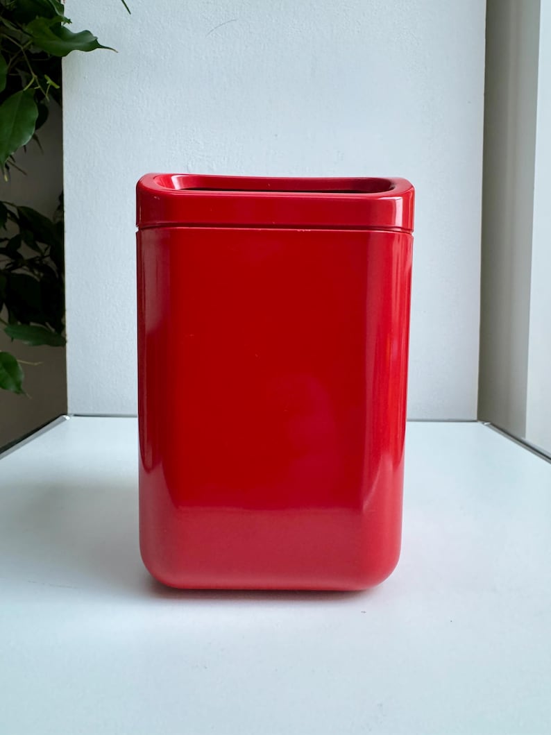 Puede incluir: Un recipiente rojo brillante de forma rectangular con una parte superior ligeramente empotrada. El recipiente tiene esquinas redondeadas y una superficie lisa y reflectante. El fondo es una superficie blanca lisa.